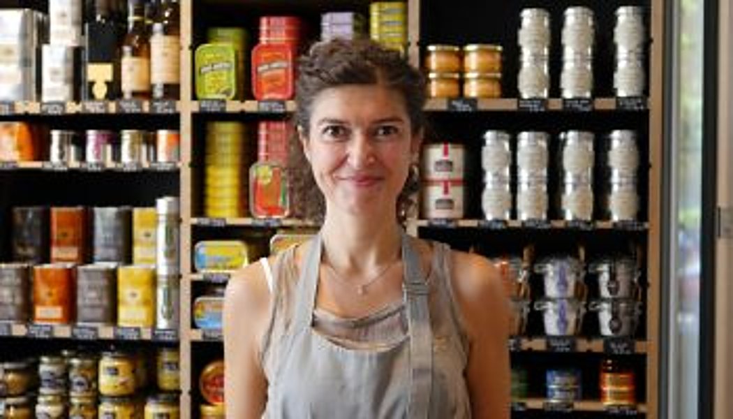 Caroline, souriante, en tablier devant les étagères de conserves de l’épicerie.