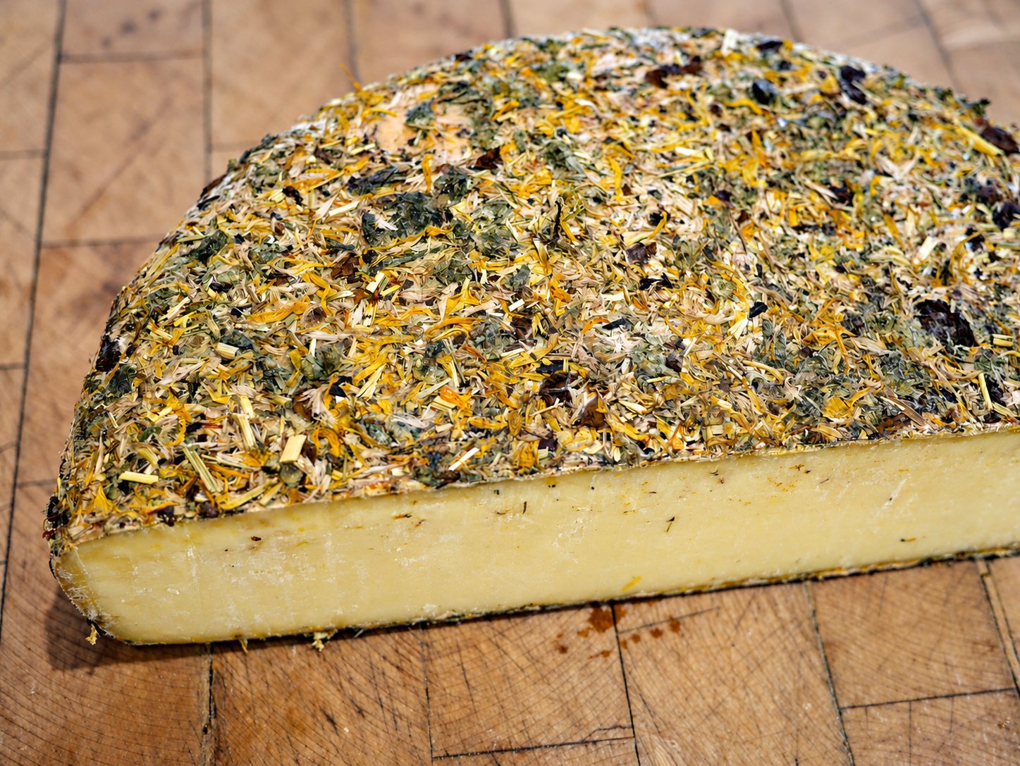 Une demi-meule de fromage à la croûte couverte d’herbes et de pétales séchés, vue de plus près.