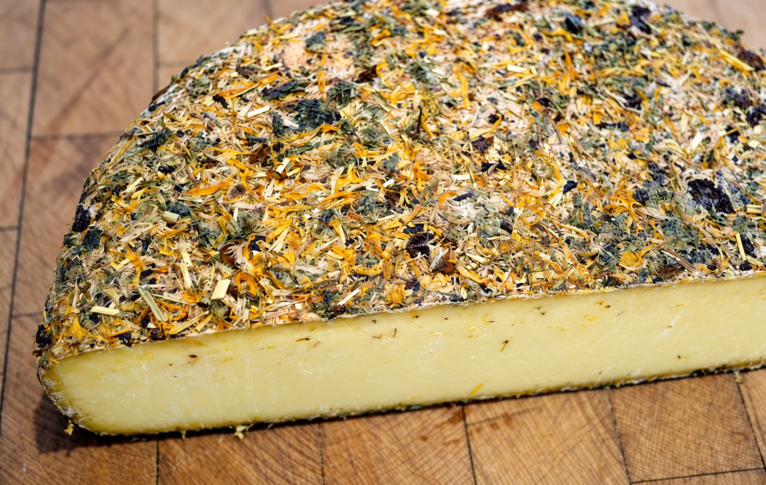 Une demi-meule de fromage à pâte pressée dont la croûte est couverte d’herbes et de pétales séchés, sur un billot de bois.