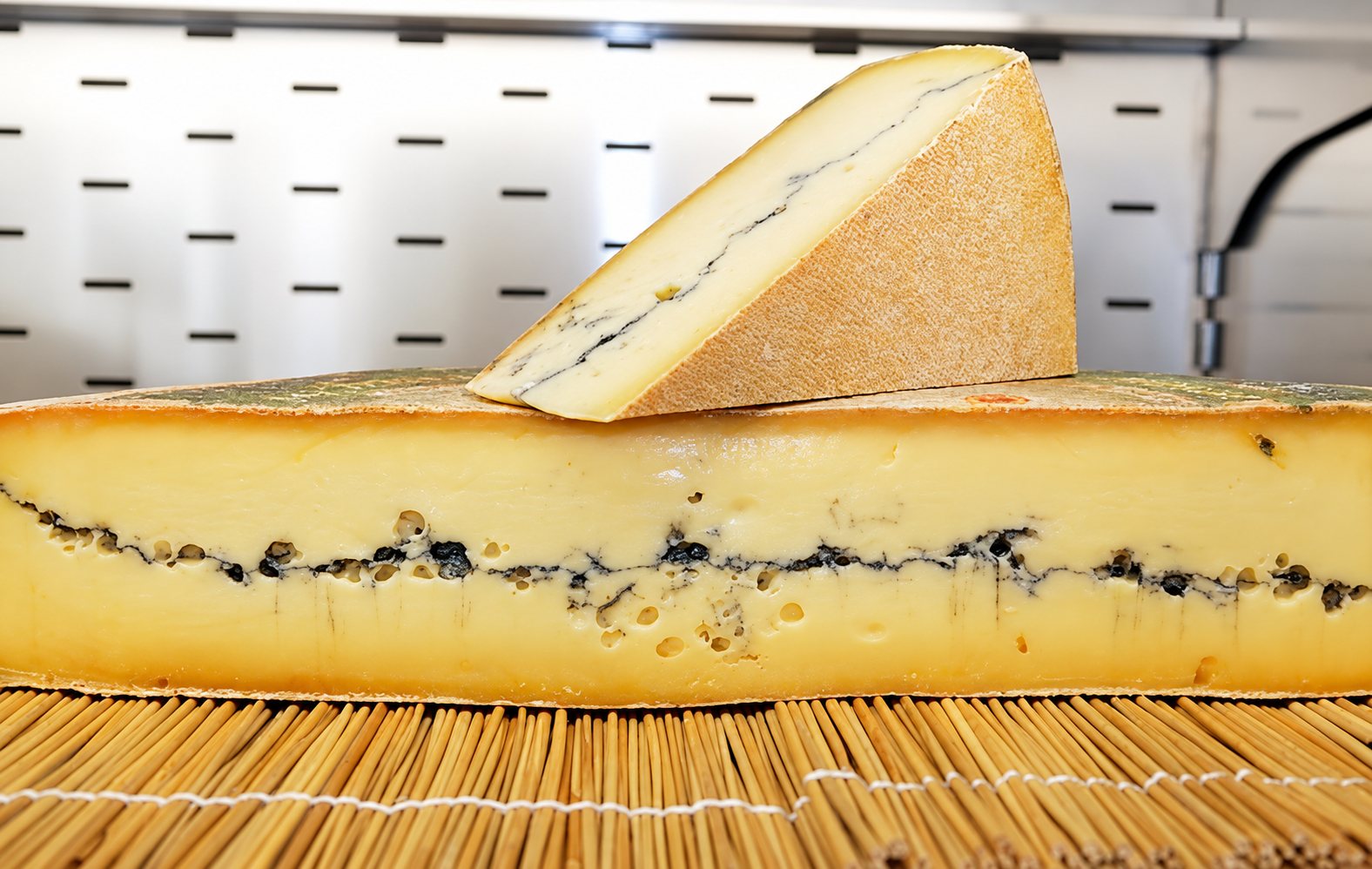 Une grande meule de Morbier avec sa raie de cendre, surmontée d’une part triangulaire, sur un tapis en bambou.