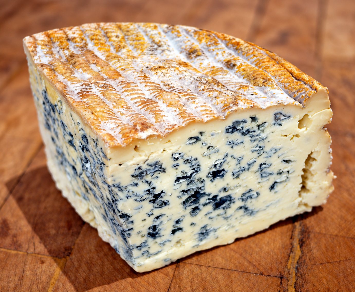 Une part triangulaire de fromage bleu à la croûte orangée et blanche, sur une planche à découper en bois.